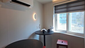 Um quarto pequeno com uma mesa redonda, um banquinho vermelho, uma janela com persianas, um ar-condicionado fixado na parede e uma luminária na parede.
