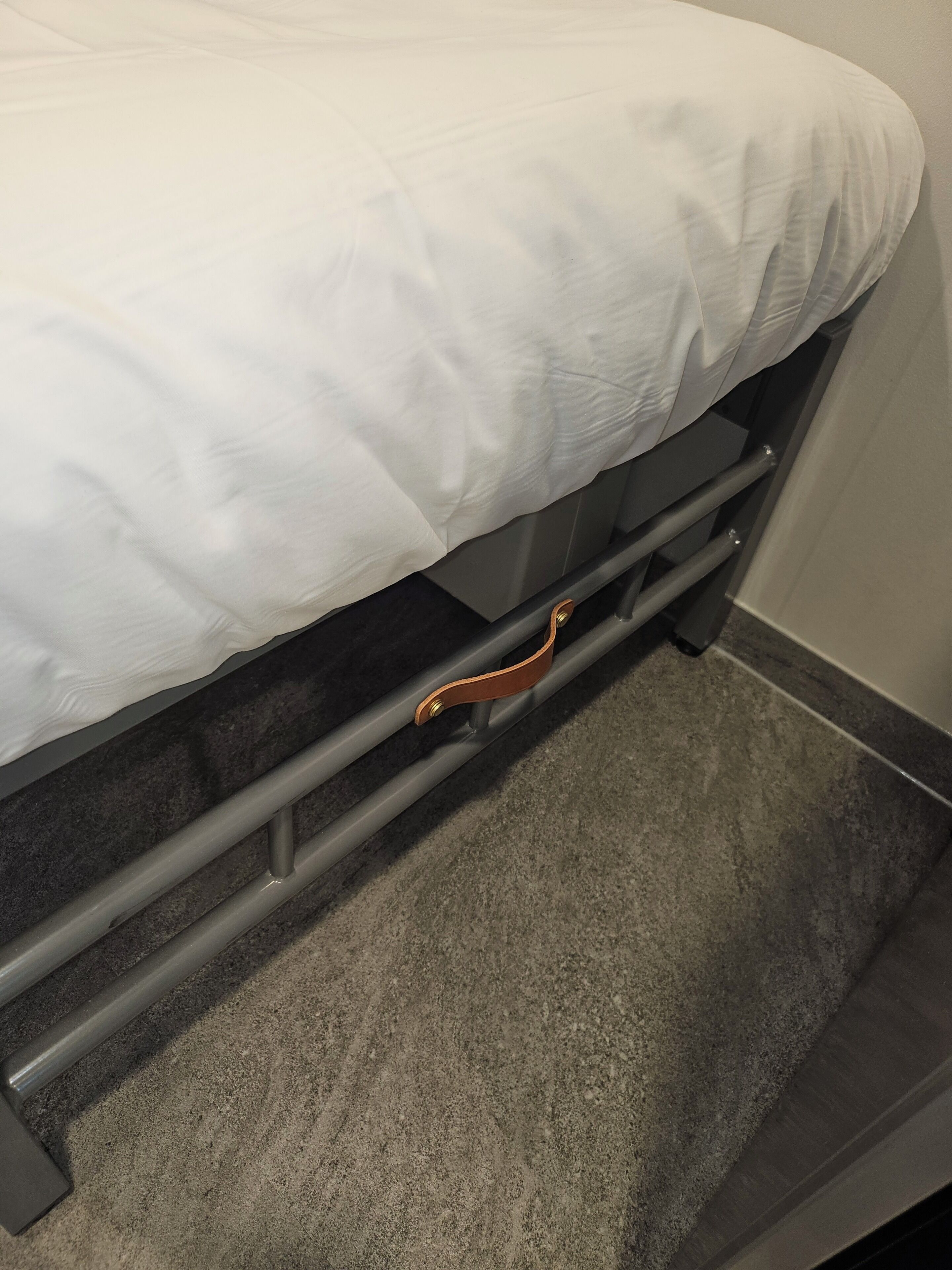 Una cama con sábana blanca, estructura de metal y un asa marrón.
