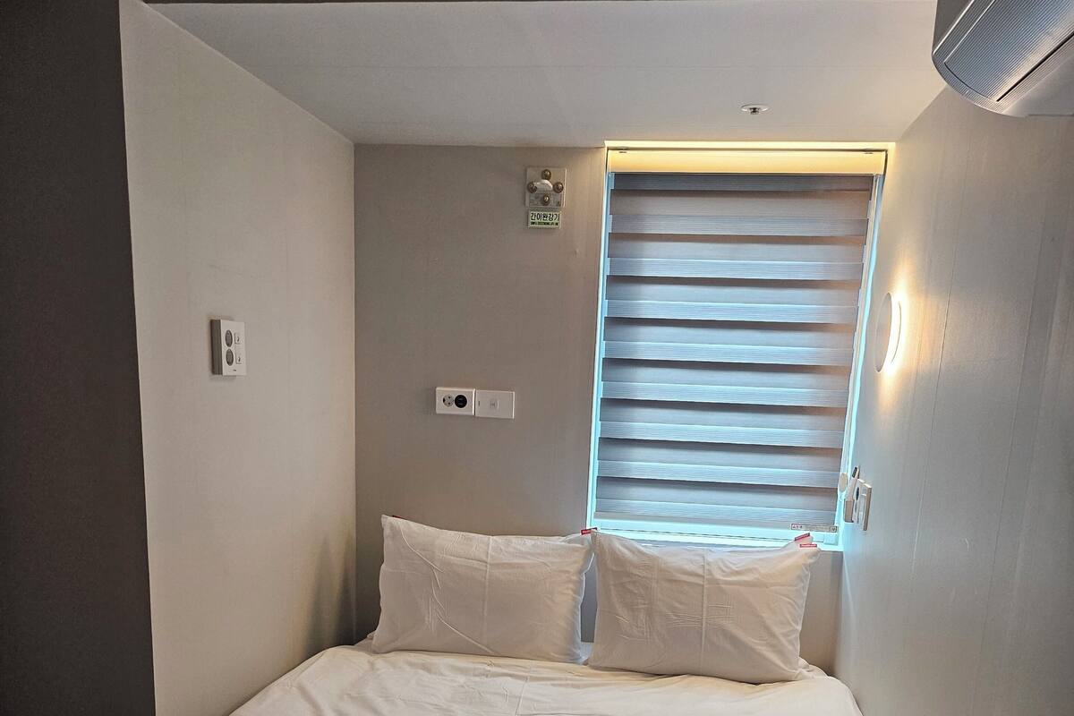 A single bed with white bedding, a window with blinds, a wall-mounted thermostat, and a ceiling light fixture.