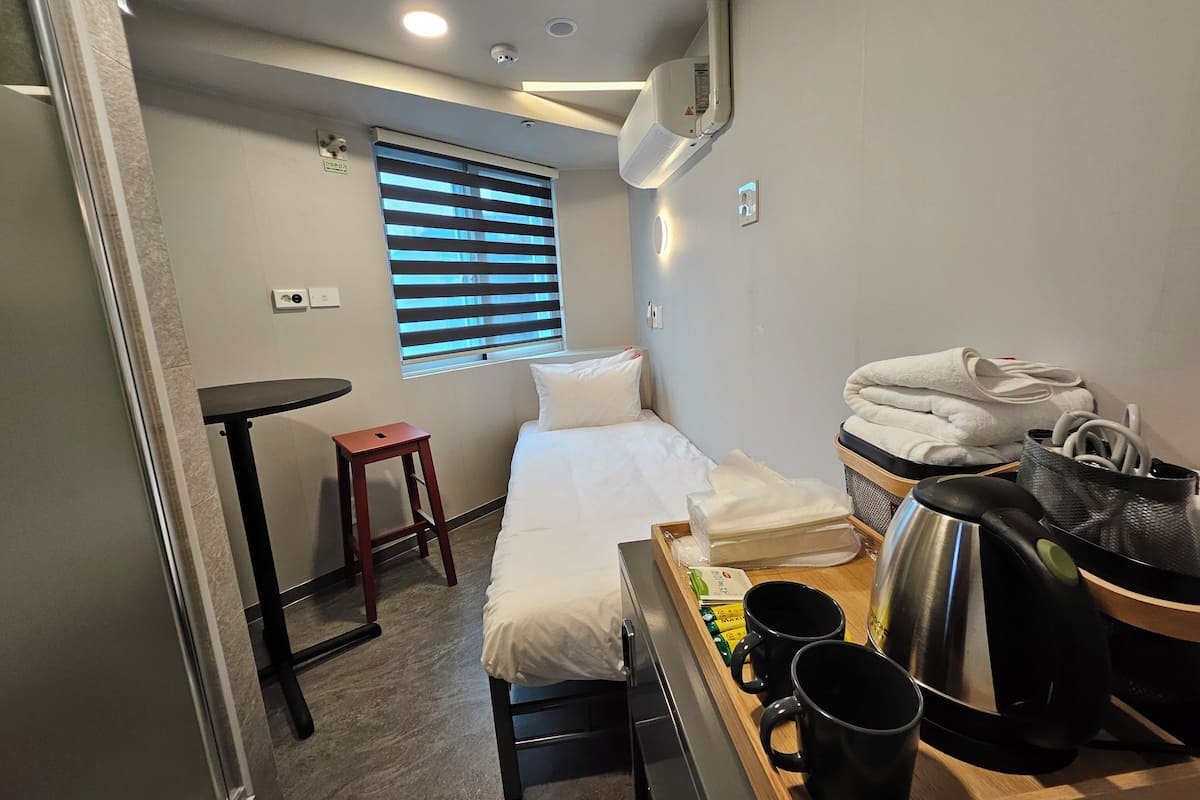 A compact hotel room with a bed, a small table, a stool, a window with blinds, a wall-mounted air conditioner, and a tray with toiletries.