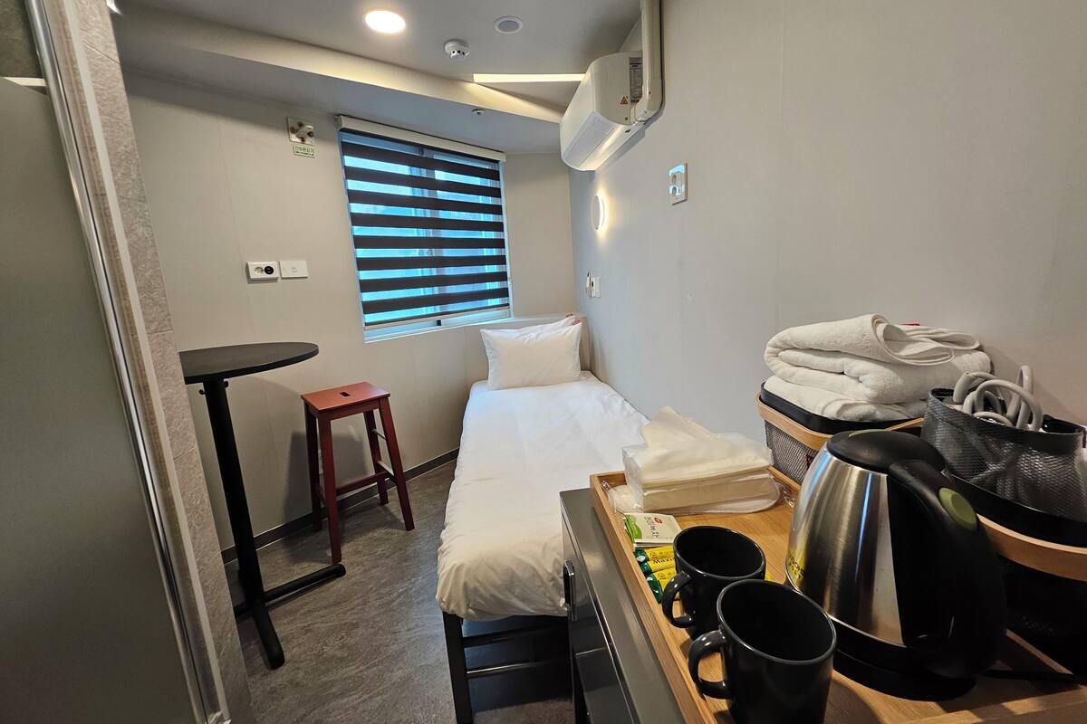 A compact hotel room with a bed, a small table, a stool, a window with blinds, a wall-mounted air conditioner, and a tray with toiletries.