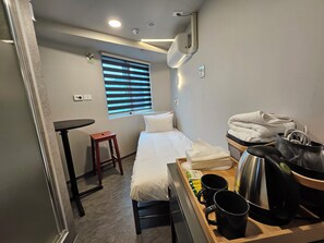 Ein kleines Hotelzimmer mit einem Bett, einem Tisch, einem Hocker, einem Fenster mit Jalousien, einer Wand-Klimaanlage und einem Tablett mit Toilettenartikeln.