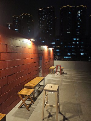 A rooftop terrace with brick walls, wooden furniture, and a view of high-rise buildings at night.