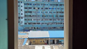 A view through a window of a multi-story building with a blue roof, surrounded by taller buildings. - RedRoof (Seoul)
