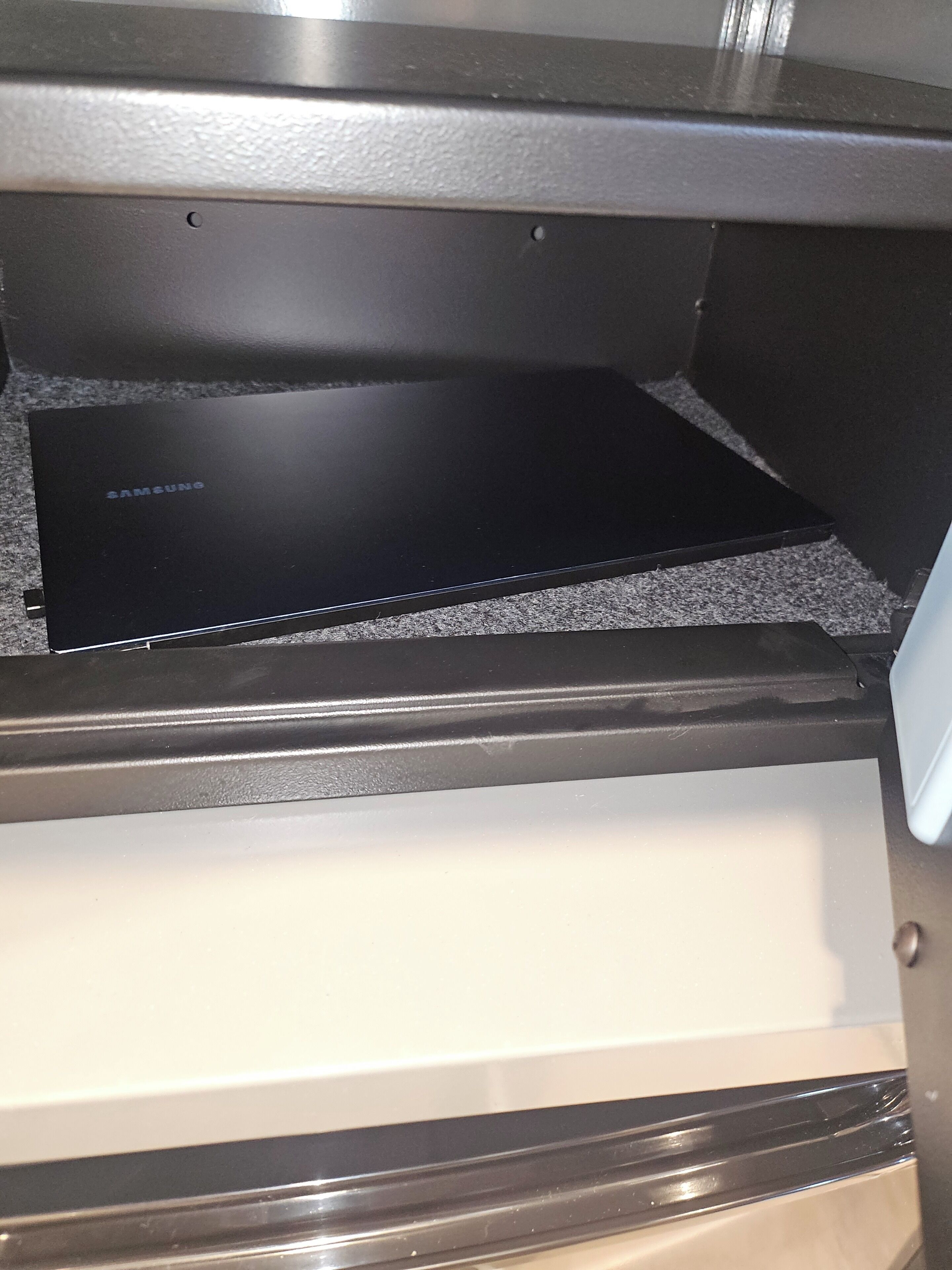 Een zwart Samsung apparaat op een gestructureerd oppervlak, een witte lade eronder en een metalen frame.