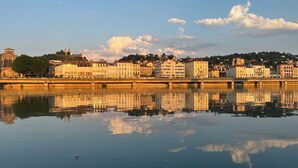 Une rivière paisible où se reflètent des bâtiments, un pont et une colline surmontée d’une tour.