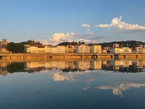 A calm river reflecting buildings, a bridge, and a hill with a tower.