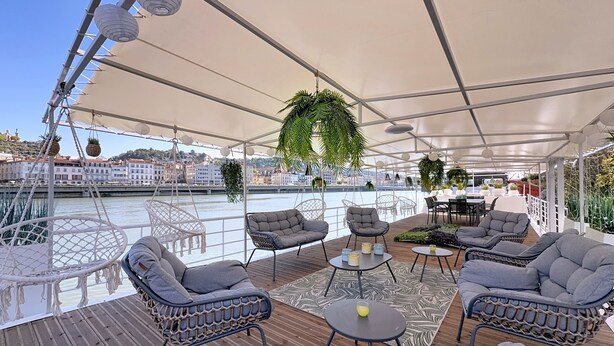 Un espace extérieur aménagé, abrité et doté de chaises en osier, de tables rondes et de plantes suspendues.
