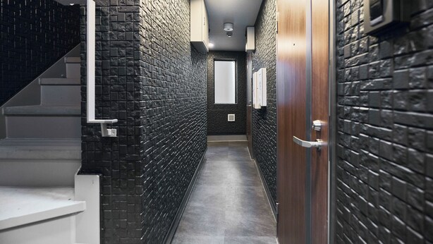 A narrow hallway with a staircase on the left, a wooden door on the right, and a wall with a brick pattern.