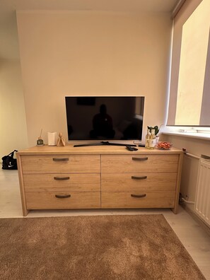 Ein Zimmer mit Fernseher auf einem Holzkasten, Heizkörper und Fenster.