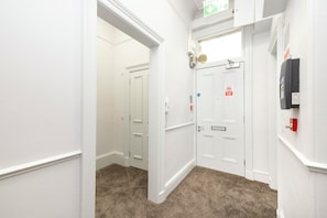 A hallway with white walls, a carpeted floor, and a door with a fire alarm. - Spacious City-Centre Home with Garden (Edinburgh)