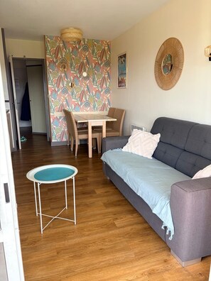 A living room with a grey sofa, a round coffee table, a dining table with wicker chairs, and a colorful floral wallpaper. - CAP ESTEREL Studio (Saint-Raphaël)
