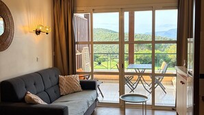 Ein Zimmer mit einer Couch, einem kleinen Tisch, einer Essecke mit Stühlen und Blick auf eine grüne Landschaft durch große Fenster.