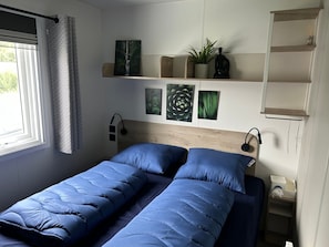 A bedroom with a bed, a bedside table, a wall-mounted shelf with decorative items, and a window with curtains. - Kamperland Beach Resort - 622 (Kamperland)