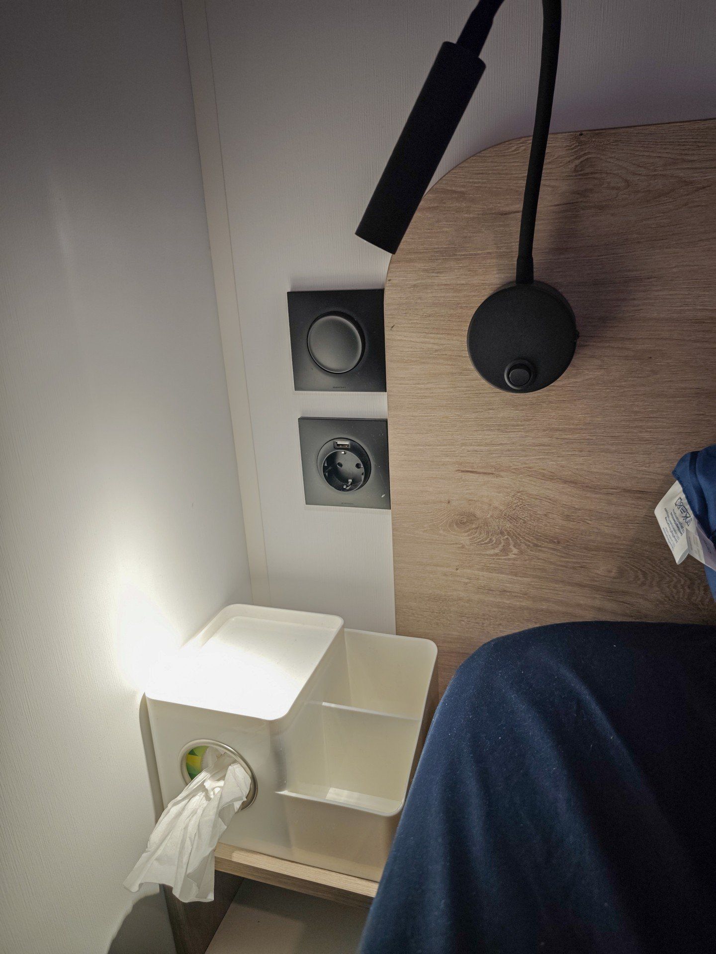 A bedroom with a wooden headboard, a bedside lamp, a white bedside table, and a wall-mounted power outlet.
