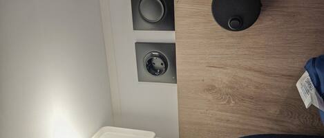 A bedroom with a wooden headboard, a bedside lamp, a white bedside table, and a wall-mounted power outlet.