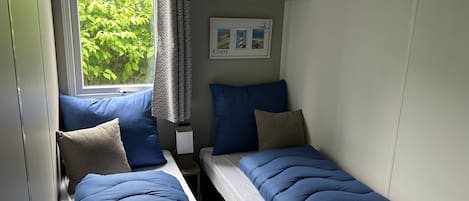 Una habitación pequeña y compacta con dos camas individuales, una ventana con vistas a zonas verdes, un estante en la pared con una planta y un cuadro con el texto 'COAST'.