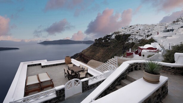 Una terrazza con poltrone e un tavolo, con vista su una località costiera caratterizzata da edifici bianchi e affaccio sul mare.