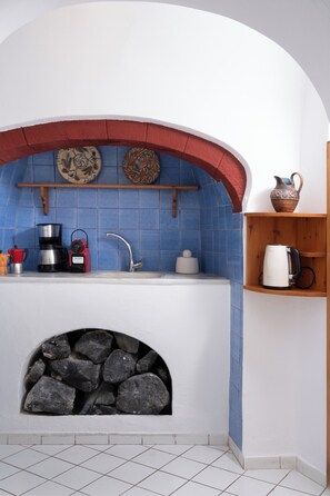 A kitchen with a stone fireplace, a sink, and various kitchen appliances. - Folia Bianca-Coco & Belle (Imerovigli)