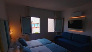 Quarto de hotel com uma cama, um sofá azul, duas janelas e uma TV fixada na parede.