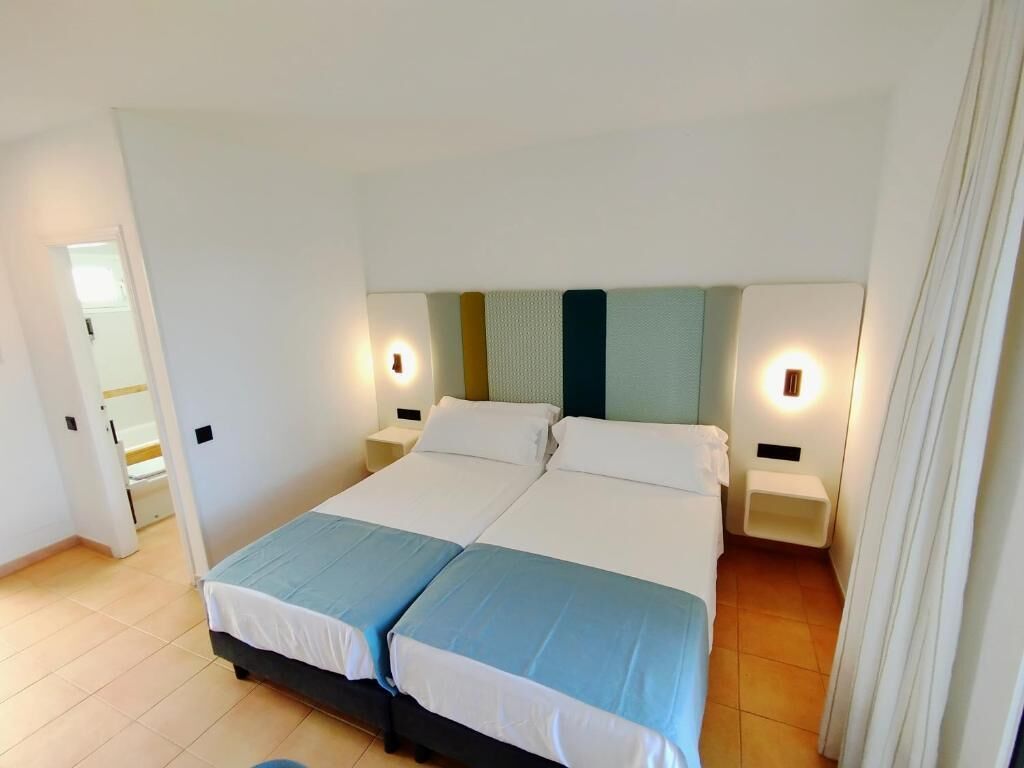 A double bed with two nightstands, a headboard with a textured panel, and a door leading to another room.