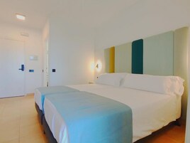 A hotel room with a large bed, a bedside table, a nightstand, a wall-mounted light, and a door.