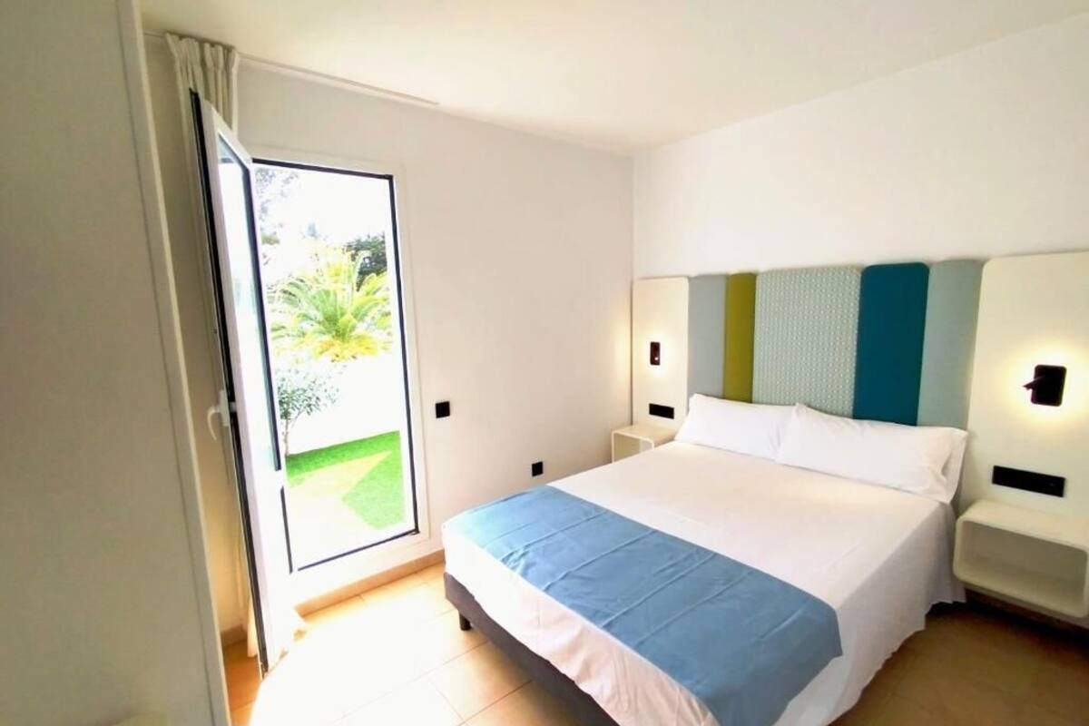 Habitación de hotel con cama, mesita de noche, lámpara, puerta corredera a un balcón y vistas a la vegetación.