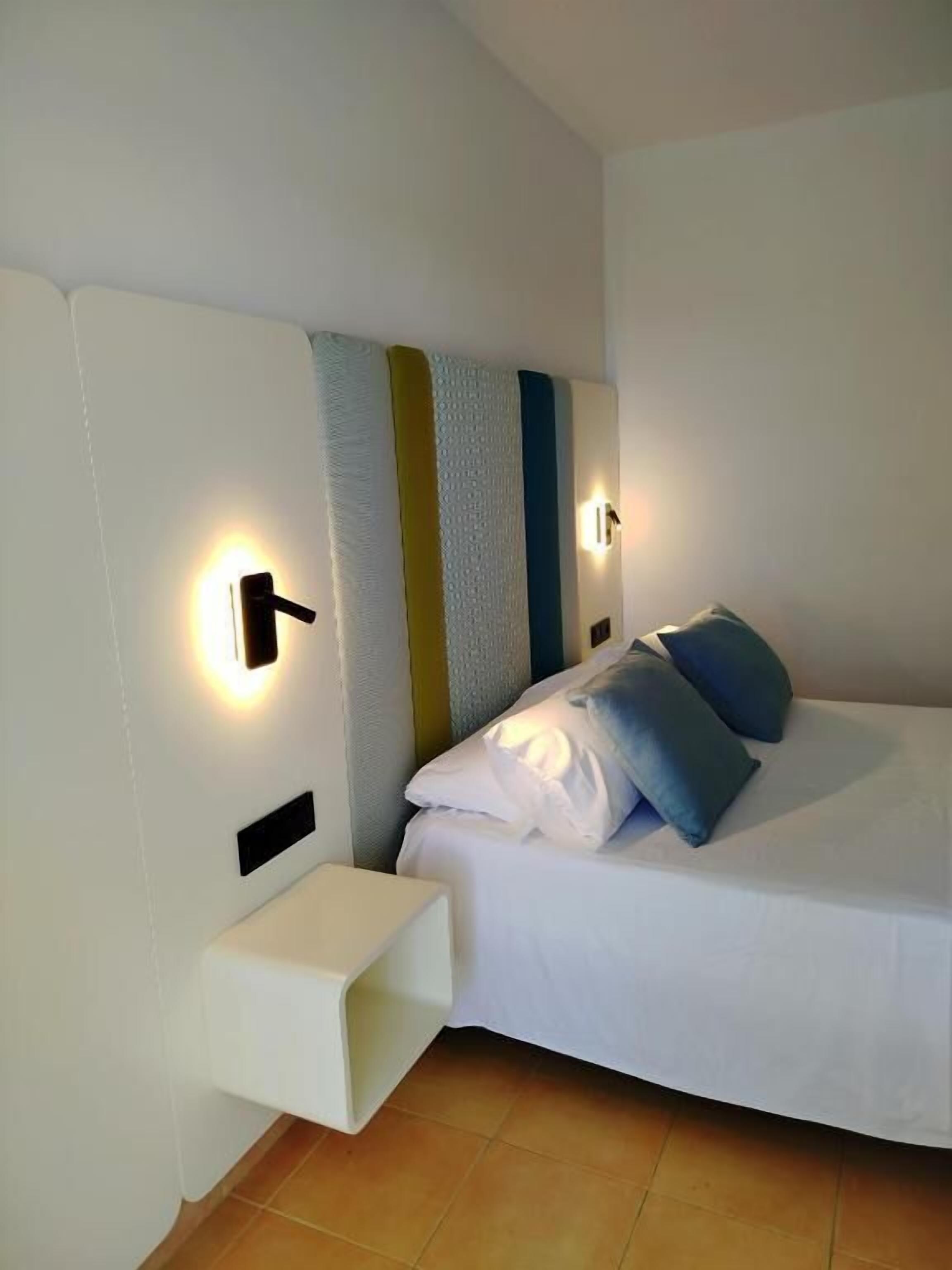 Un dormitorio moderno con cama, lámparas de noche y una mesita de noche pequeña y blanca.