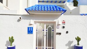Entrada de um edifício com porta azul, uma placa com a letra 'H' e dois cactos em vasos.