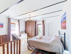 Une chambre à coucher comprenant un lit, une chaise, une petite table, une armoire en bois et une fenêtre avec des rideaux.