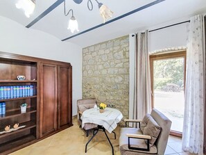 Ein Zimmer mit einer Steinswand, einem hölzernen Bücherregal, einem Tisch mit weißem Tischtuch, einem Sessel und Blick durch ein Fenster mit Vorhängen.