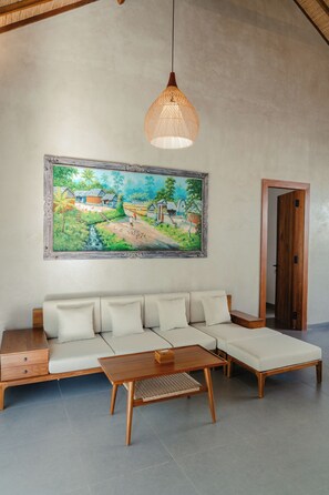 Ein Wohnzimmer mit einem Ecksofa, einem Holztisch, einer hängenden Pendelleuchte und einem großen Landschaftsbild an der Wand.