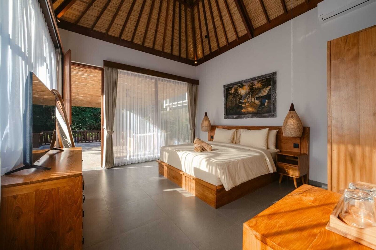 Ein Schlafzimmer mit einem großen Bett, einem Holzkopfende, einem Bild an der Wand und Blick durch das Fenster.