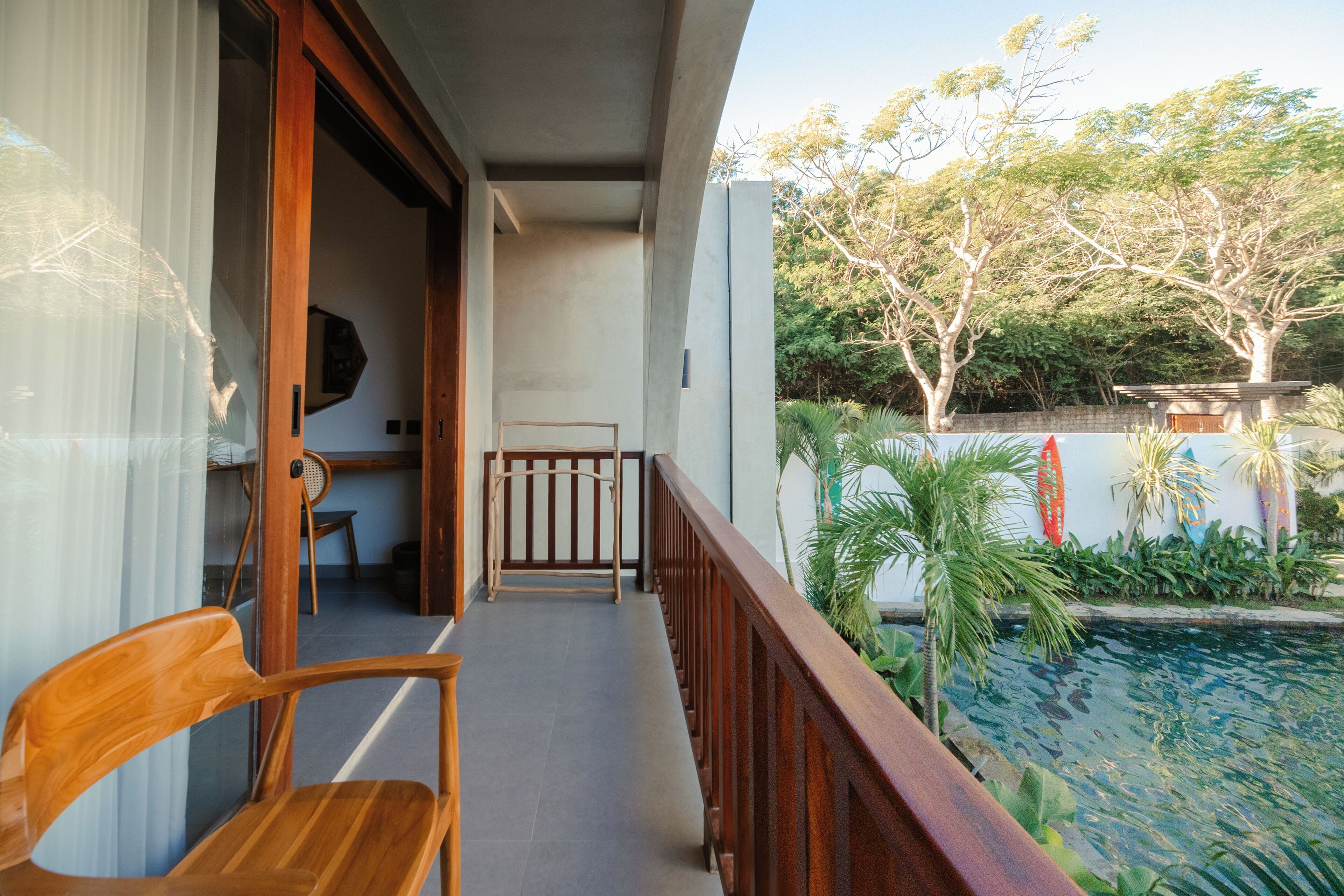 Un balcón con una silla de madera, una mesita y vistas a una piscina y vegetación.