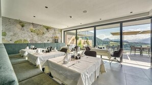 Un restaurant avec des tables dressées pour le service, un mur de pierre agrémenté de verdure et une vue sur les montagnes, visible à travers de grandes fenêtres.