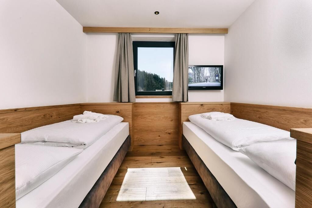 A room with two beds, a window with curtains, a flat-screen TV on the wall, and a wooden floor.