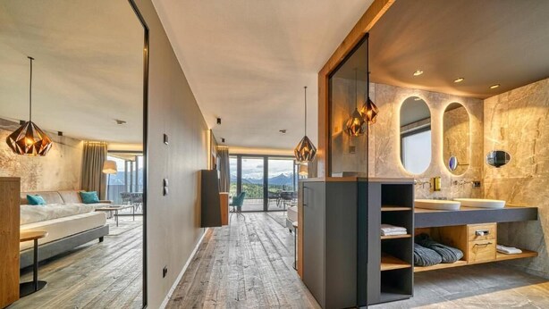Une chambre d’hôtel moderne avec un plancher en bois, un canapé, un coin repas, une grande fenêtre et une salle de bain avec un mur en pierre et un lavabo.