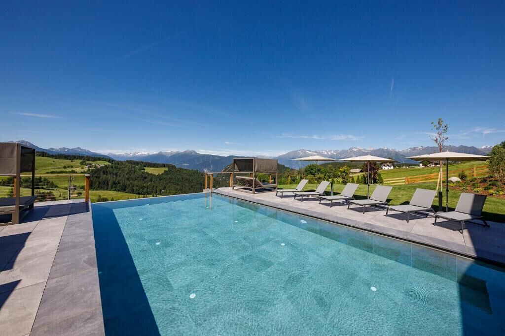 Piscina con sillones de descanso y sombrillas, rodeada de montañas y cielos despejados.