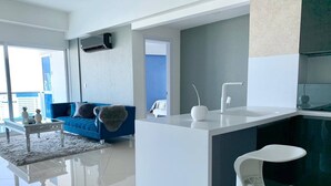 Ein modernes Wohnzimmer mit einem blauen Sofa, einem weißen Couchtisch und einer Küchenzeile mit weißer Arbeitsplatte und einem weißen Stuhl.
