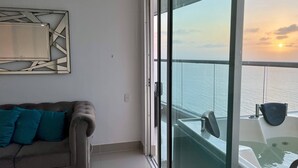 Een balkon met uitzicht op zee bij zonsondergang, een bank met turquoise kussens en een modern abstract kunstwerk aan de muur.