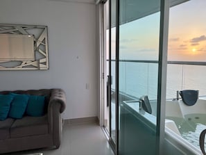 Ein Balkon mit Meerblick bei Sonnenuntergang, eine Sitzecke mit türkisfarbenen Kissen und ein modernes, abstraktes Wandbild.