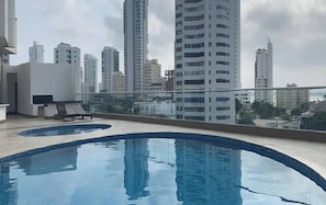 Ein Pool auf dem Dach mit Glasbrüstung, eine Lounge mit Flachbildfernseher und hohe Gebäude im Hintergrund.