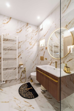 A bathroom with a white sink, gold fixtures, a marble wall, a white towel rack, and a round mirror.