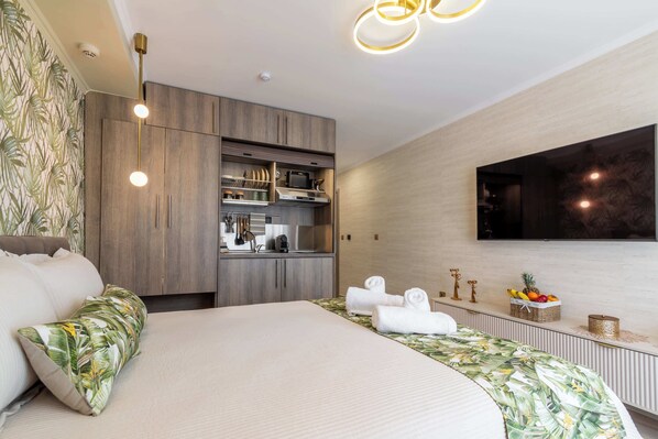 Habitación de hotel moderna con cama, kitchenette, televisión y estante en la pared con toallas y fruta.