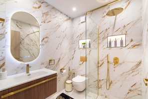 A modern bathroom with a marble wall, a round mirror, a gold faucet, a toilet, a shower, and a small shelf with toiletries.