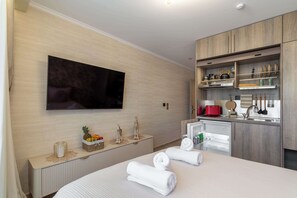 Ein modernes Hotelzimmer mit Flachbildfernseher, einem Bett mit ordentlich gefalteten Handtüchern, einer kleinen Küchenzeile und einem an der Wand montierten Fernseher.