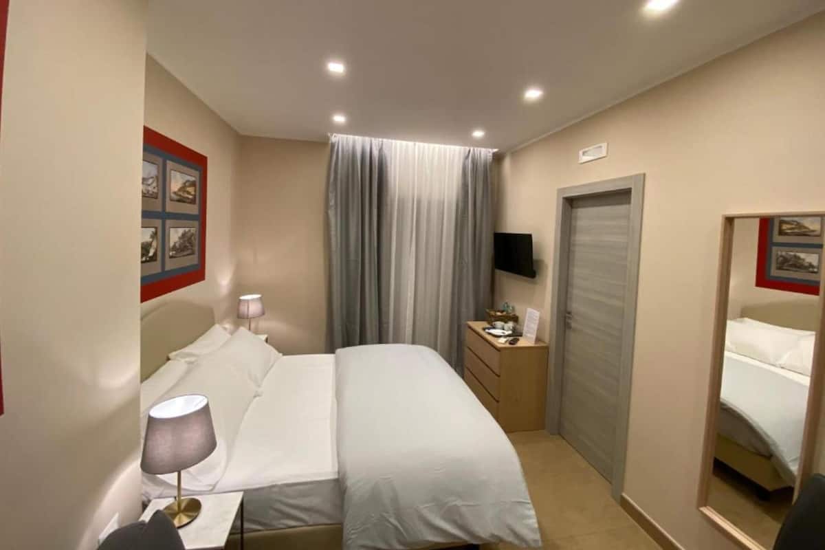 Et hotelværelse med seng, sengebord, lampe, gardiner, et fjernsyn og et skab.