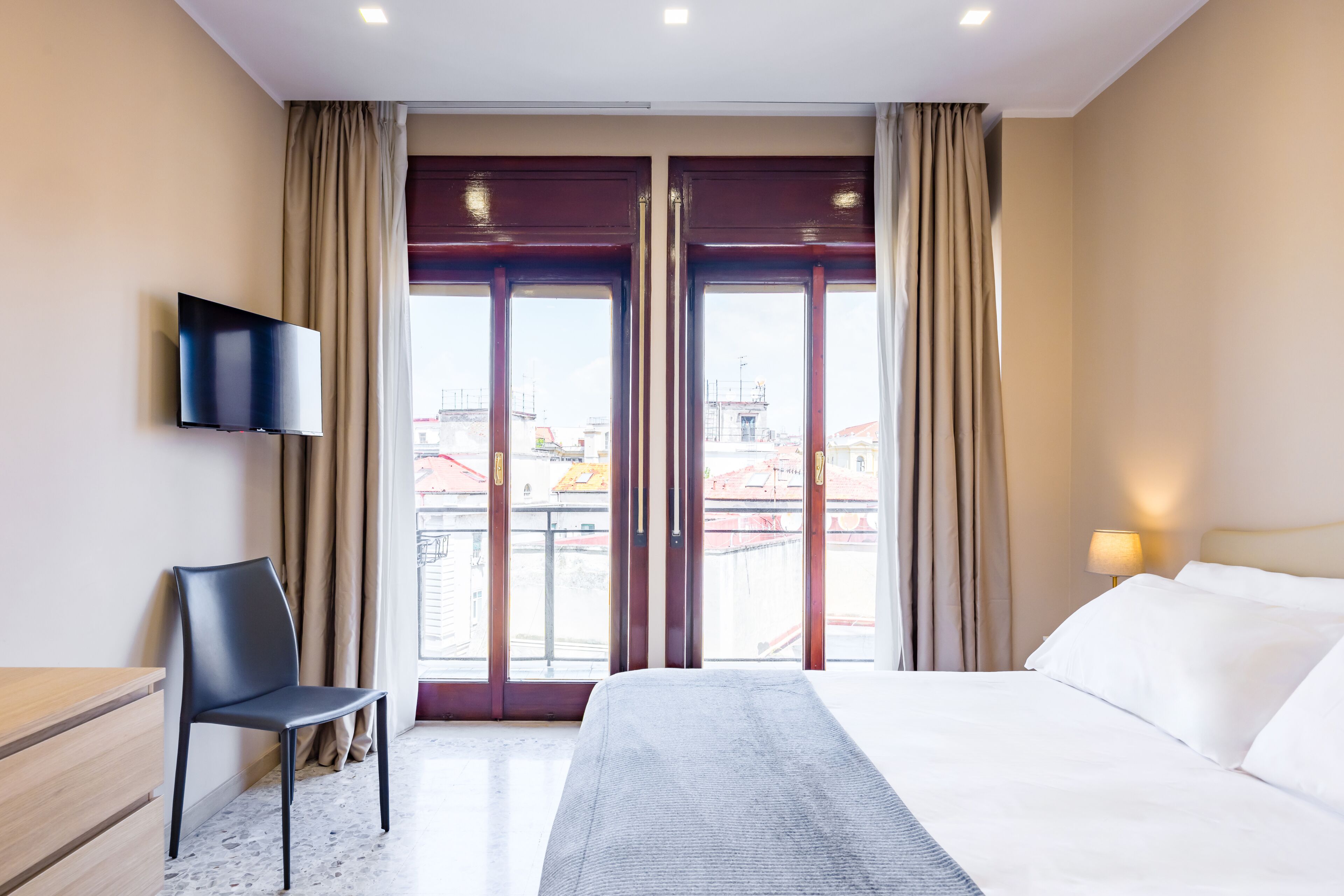 A hotel room with a bed, a chair, a TV, and a view of buildings through the window.
