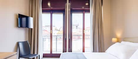 A hotel room with a bed, a chair, a TV, and a view of buildings through the window.