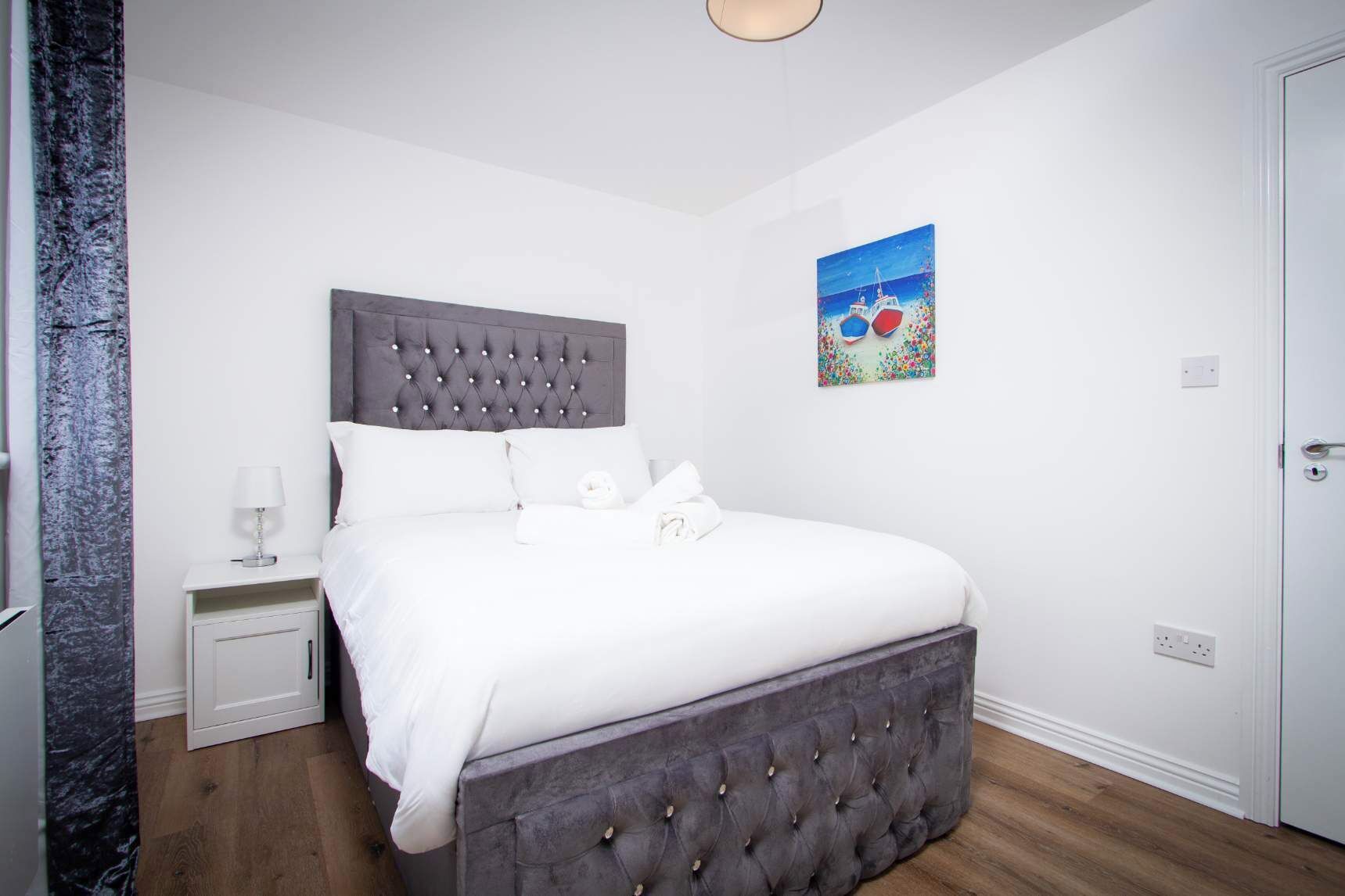 Un dormitorio con cabecera capitonada, ropa de cama blanca, una mesita de noche con lámpara y un cuadro en la pared.